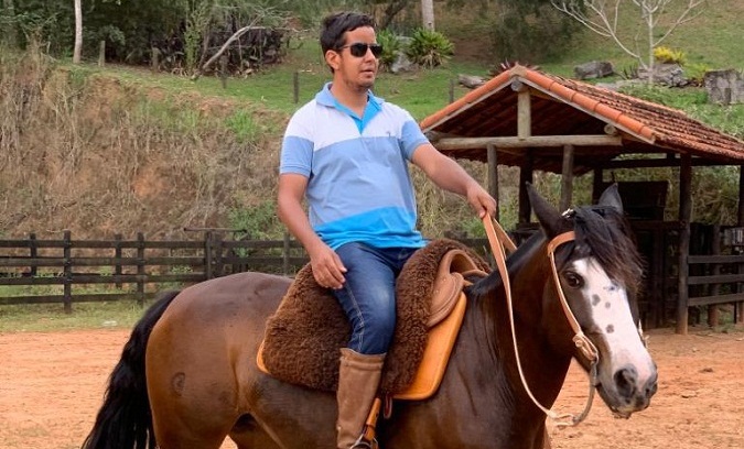 Na imagem Hugo Antônio está montado em um cavalo de cor marrom. Ele está vestido com uma uma blusa de manga curta, na cor azul, uma calça jeans azul e calçado com uma bota. 